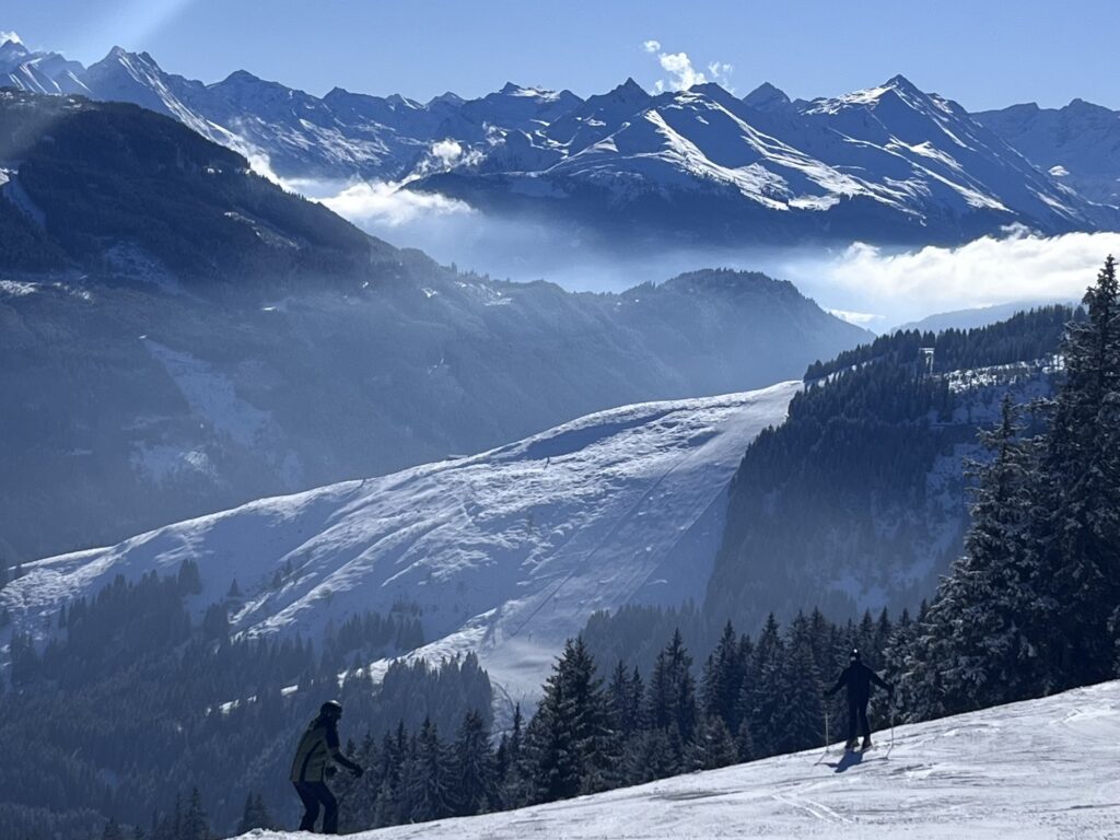 Zwei Skifahrer fahren auf einer sonnigen Piste in KitzSki und genießen den beeindruckenden Blick auf die verschneiten Gipfel der Kitzbüheler Alpen, die sich über einem Nebelmeer erheben.